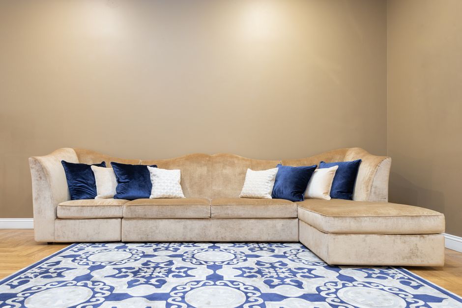 Close-up view of a large, cushioned sofa with blue velvet upholstery and decorative pillows, situated in a well-lit living room. The sofa features an elegant curved armrest with a dark, ribbed finish 