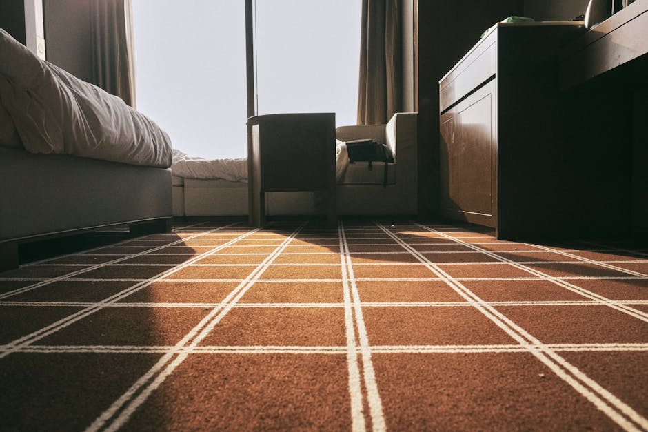 A person kneeling on a patterned carpet in a residential setting, preparing a yellow portable vacuum cleaner with a black hose for surface cleaning. The individual, dressed in casual clothing, is open